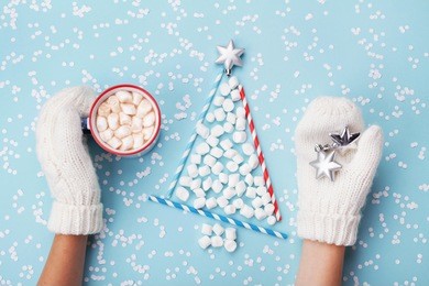 woman hand in mitten hold cup of hot cocoa or chocolate and christmas fir tree made of marshmallow on turquoise winter background top view. flat lay. holiday celebration concept.