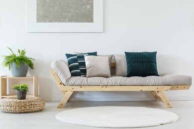 plant on pouf near beige settee with pillows in living room with white round carpet and silver painting