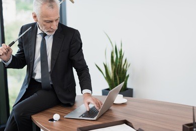 a respectable elderly man in a strict business suit is posing in his office with a golf club. he has a serious face