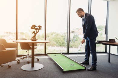 a man in a business suit plays mini golf. he is in a modern modern office