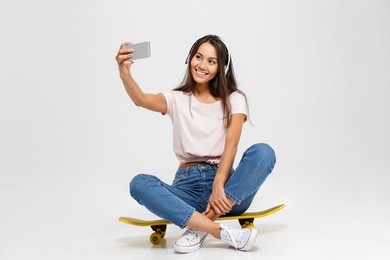 portrait of young cheerful girl in white headphone makes selfie on smartphone, sitting on yellow skateboard, isolated over white background