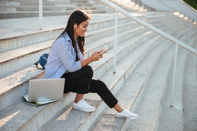 portrait of beautiful asian student, listening to music, holding mobile phone, sitting on the city stairs
