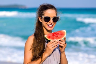 smiling woman with long shiny hair posing at sea in sunny summer day. outdoor portrait of stylish tanned girl in sunglasses eating watermelon and walking down the beach.