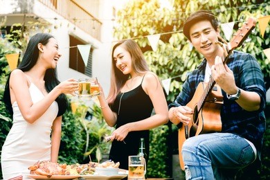asian young women enjoying toasting drinks party with guy playing guitar singing at home garden outdoors.