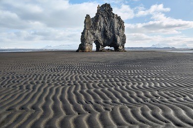 abstract rock on the dark volcanic sand on the background of the blue sky with clouds. it is hvitserkur during low tide in iceland. horizontal.