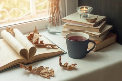 autumn still life. books, leaves, cup and dry tiny flowers in vase red candlestick on window. concept of autumn reading time and romantic, warm, cozy seat  opened book, rustic style home decor