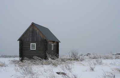 the small wooden house on the snow-covered field with dried grass on the cloudy winter day. 