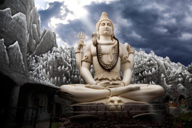big lord shiva statue sitting in lotus with trident in his hand and cobra near by. dramatic sky at background with ray on shiva. bangalore, india
