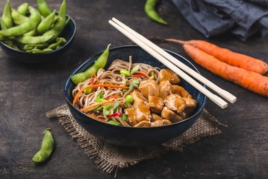 asian noodles with chicken, vegetables in bowl, rustic wooden background. soba noodles, teriyaki sauce chicken, edamame beans, sesame, chopsticks. closeup. asian style dinner. chinese/japanese noodles