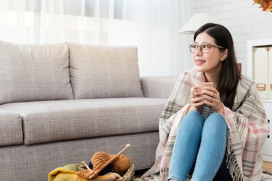 smile asian woman drinking hot chocolate and keeping warm with the wool blanket and looking outside autumn view and knitting on the floor in the living room at home