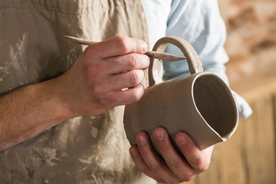 pottery, workshop, ceramics art concept - closeup on young ceramist hands with unbaked clay jug, craftsman stand at a workplace, hands holds the clay cup and tools