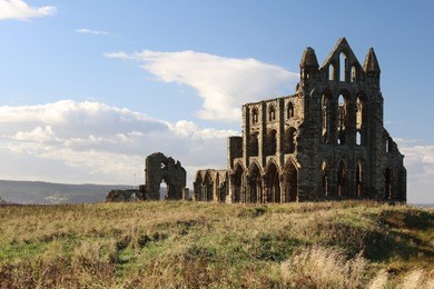 a wide shot of whitby abbey ruins in evening sunlight. there is blue sky behind the abbey and grassland in front. the abbey itself is part in sun part in shade.