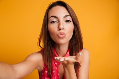pretty brunette woman in dress making selfie and sends air kiss at the camera over yellow background