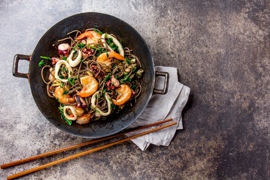 seafood noodles. buckwheat stir-fry noodles with seafood - shrimps, octopus, squid in cast iron asian wok with cooking chopstick. top view, stone background. 