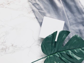 white plain square box or package next to a big tropical leaf and grey scarf on white marble desk background. copy space. flat lay. top view