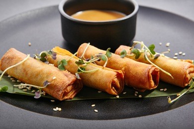 spring rolls - fried vietnamese rolls of rice edible paper on black matte plate next to cup with sauce. pan-asian menu of restaurant.