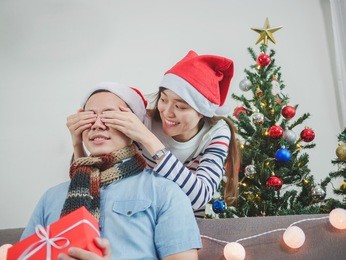 asian young woman giving a christmas present to his boyfriend on sofa at home, have christmas tree ornaments.