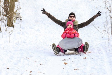 beautiful sporty caucasian girl with daughter having fun and sledding in a forest or city park on a bright sunny day. winter family activity concept.
