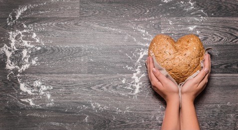 hands holding heart shaped homemade wheat bread on dark wooden table with some flour - top view horizontal banner copy space for text