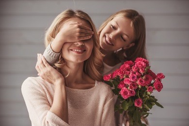 pretty teenage daughter is holding flowers and covering her mom's eyes while making a surprise, both are smiling
