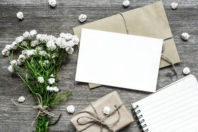 blank white greeting card with white flowers bouquet and envelope with gift box and paper notebook on rustic wooden background .top view. mock up