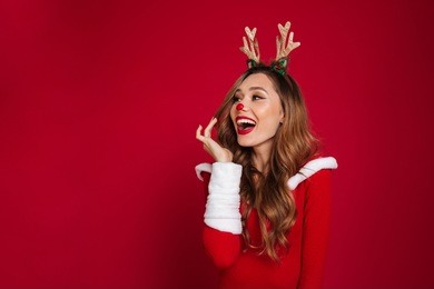 portrait of an cheerful happy girl wearing christmas deer costume while standing and looking away at copy space isolated over red background
