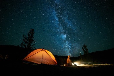 tourist near his camp tent at night under a sky full of stars. orange illuminated tent.