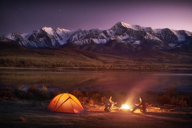 night camping. two men tourists sitting at the illuminated tent near campfire under amazing sunset evening sky in a mountains area. snow mountain in the background