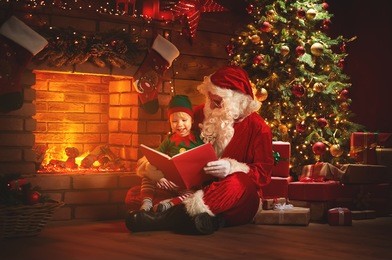 santa claus reads a book to a little elf by fireplace and christmas tree
