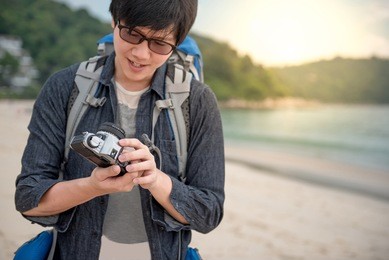 young asian man traveler look at his vintage camera, happy male photographer on the beach, summer holiday and vacation lifestyle concepts