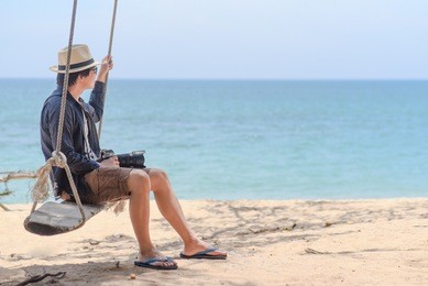 young asian photographer man on wooden swing at the beach, male traveler relaxing with seascape in summer season of thailand, travel lifestyle concept