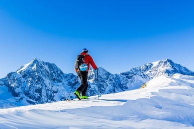 mountaineer backcountry ski walking up along a snowy ridge with skis in the backpack. in background blue sky and shiny sun and zebru, ortler in south tirol, italy.  adventure winter extreme sport. 