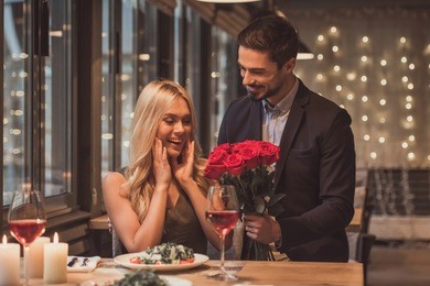 handsome elegant man is giving roses to his beautiful girlfriend while making a surprise in restaurant, both are smiling