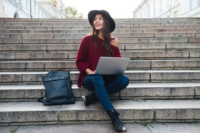 portrait of a smiling young asian woman in hat typing on laptop computer while sitting on staircase outdoors
