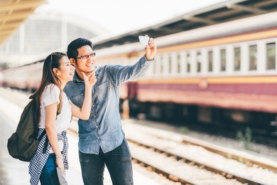 young asian couple traveler taking selfie together using smartphone waiting for trip at train station platform in asia. backpack travel, love relationship, holiday vacation or modern lifestyle concept