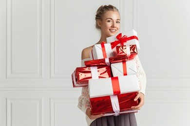 blond hair woman in white sweater holding a lot of presents over white wall background indoor. smiling woman holding paper gift boxes red color and looks at camera. toothy smiling woman's face.