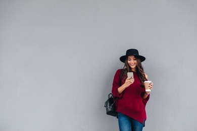 portrait of a happy asian girl dressed in hat and sweater holding coffee cup while standing and using mobile phone on a city street
