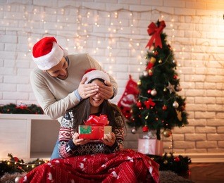 young handsome man with santa hat holding eye closed of his girlfriend while she sitting on a carpet with a present in hands for christmas holidays.