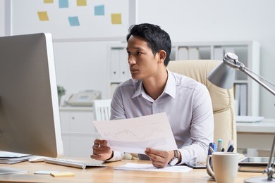 financial analyst with document in his hands reading information on computer screen