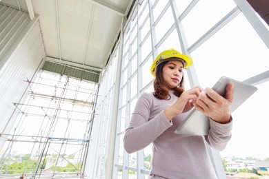 female engineer in yellow safety helmet is looking to tablet computer while checking for under construction building and curtain wall frame with scaffold in background