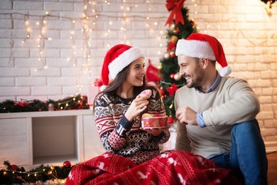 positive charming love couple sitting on a carpet with santa hats and red blanket while eating christmas cookies and looking each other with happiness.