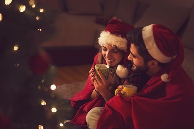 couple in love sitting next to a christmas tree, wearing santa's hats, drinking hot tea and enjoying romantic moments together. focus on the girl