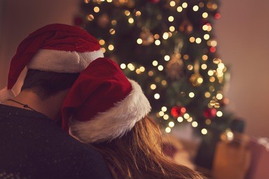 couple in love sitting next to a christmas tree, wearing santa's hats, hugging and looking away from the camera towards the tree. selective focus on the girl's hat