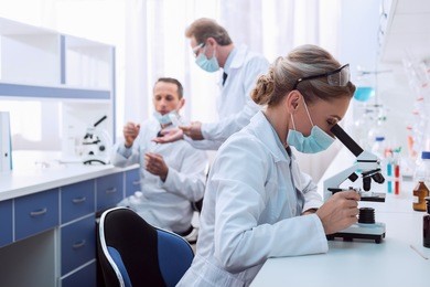 medical worker in lab coat and sterile mask, doing a microscope analysis while her colleague are working behind