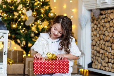 a brunette gilr in front of fur-tree and fireplace with candles and gifts. a surprised girl. a girl dreaming. new year's eve. christmas eve. cozy holiday at the fur-tree with lights and gold decor.