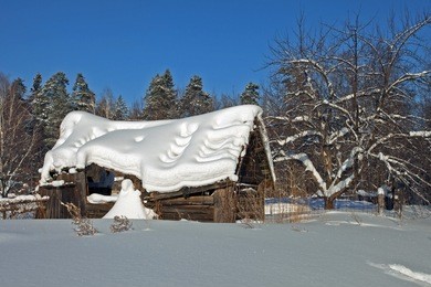 winter in the forest, a wooden house is covered with a thick layer of snow against the clear blue sky