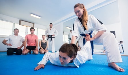 young female coach demonstrating painful hold to adults in taekwondo class
