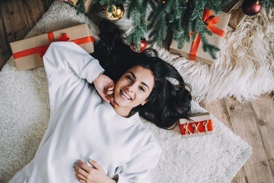 incredible and stunning young woman is lying on the white carpet between christmas gifts and beautiful green pine tree. she is relaxing and enjoying her time spanding like that. close up. cut view