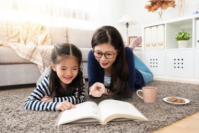 charming asian mother and her daughter reading book and enjoy hot chocolate and coffee sitting on floor of the living room at home