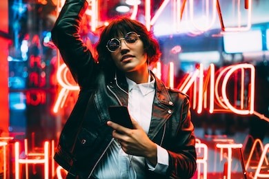 beautiful brunette woman in trendy apparel and eyewear enjoying nightlife in city listening music in earphones and moving to sound, gorgeous hipster girl dancing outdoors on neon city illumination 